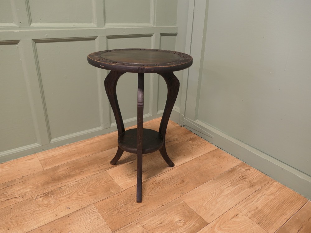 A dark oak circular Arts and Crafts lamp table featuring carved sunflower motifs around the rim, curved legs, and a lower circular tier.