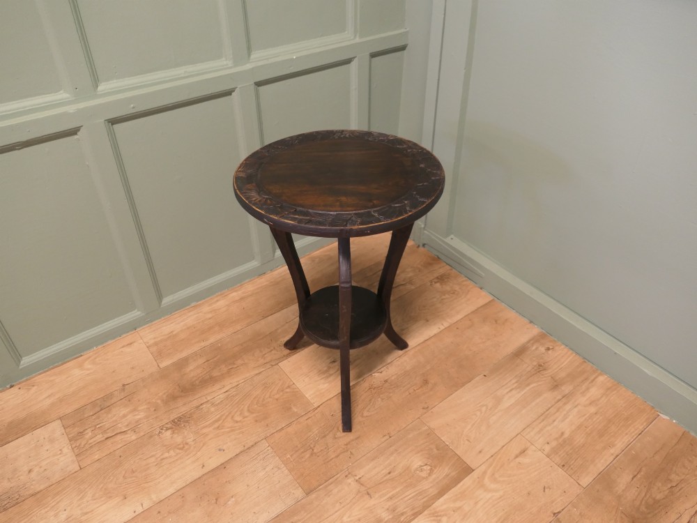A dark oak circular Arts and Crafts lamp table featuring carved sunflower motifs around the rim, curved legs, and a lower circular tier. View II