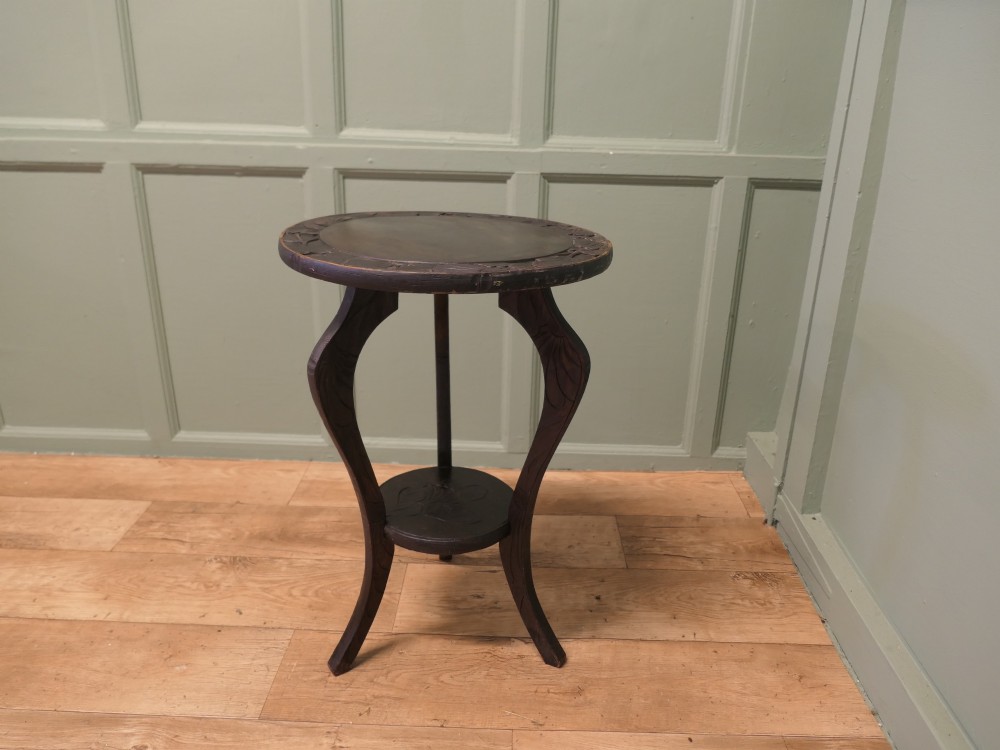 A dark oak circular Arts and Crafts lamp table featuring carved sunflower motifs around the rim, curved legs, and a lower circular tier. View III