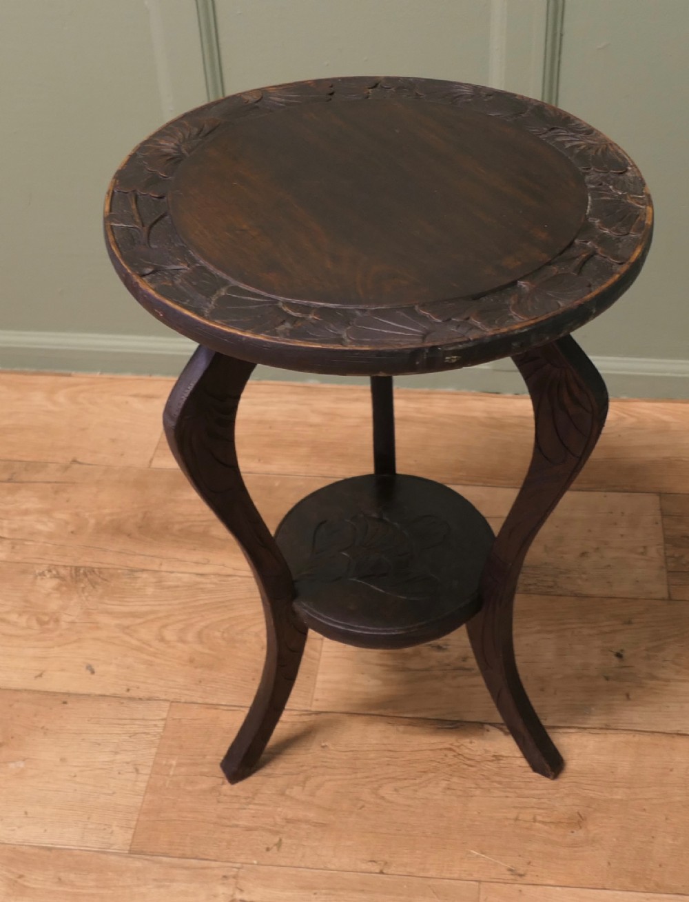 A dark oak circular Arts and Crafts lamp table featuring carved sunflower motifs around the rim, curved legs, and a lower circular tier. View IV