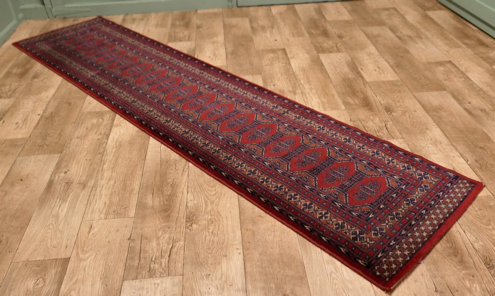 A long wool hallway runner carpet with repeating red geometric hexagonal medallions surrounded by patterned navy blue and terracotta borders. View II