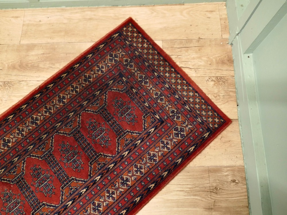A long wool hallway runner carpet with repeating red geometric hexagonal medallions surrounded by patterned navy blue and terracotta borders. View III
