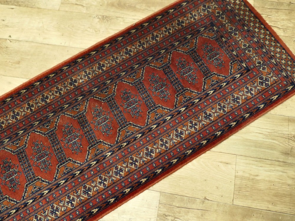 A long wool hallway runner carpet with repeating red geometric hexagonal medallions surrounded by patterned navy blue and terracotta borders. View IV