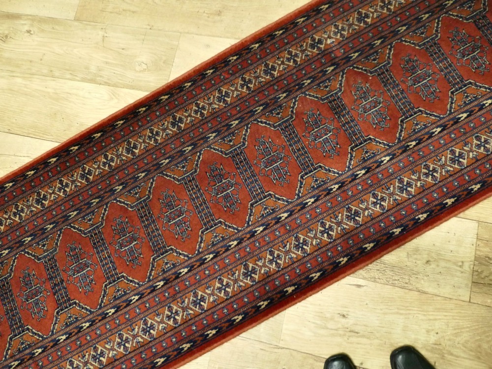 A long wool hallway runner carpet with repeating red geometric hexagonal medallions surrounded by patterned navy blue and terracotta borders. View V