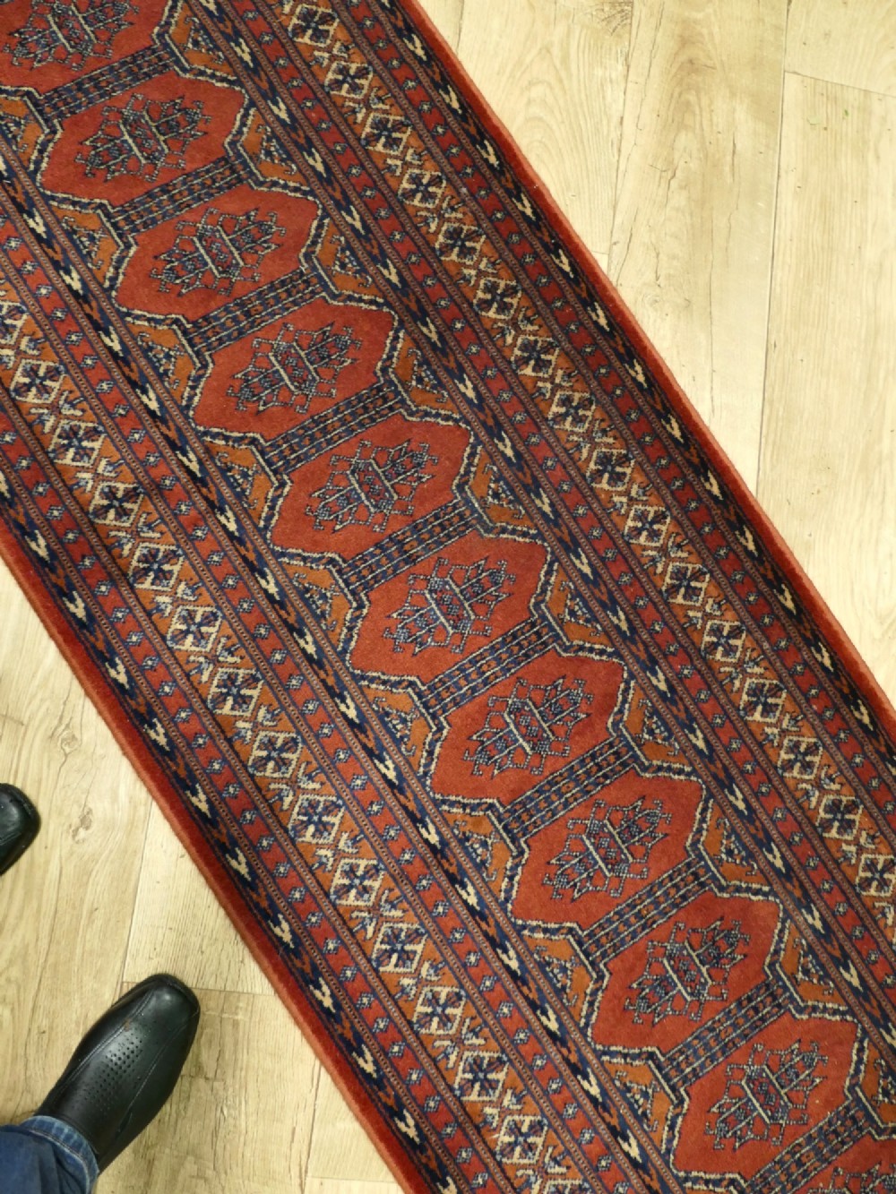 A long wool hallway runner carpet with repeating red geometric hexagonal medallions surrounded by patterned navy blue and terracotta borders. View VI