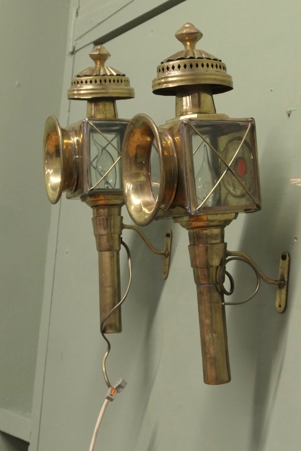 A pair of electrified brass French carriage lanterns mounted on a wall, featuring circular front glass lenses, top finials, and tubular shafts. View VI