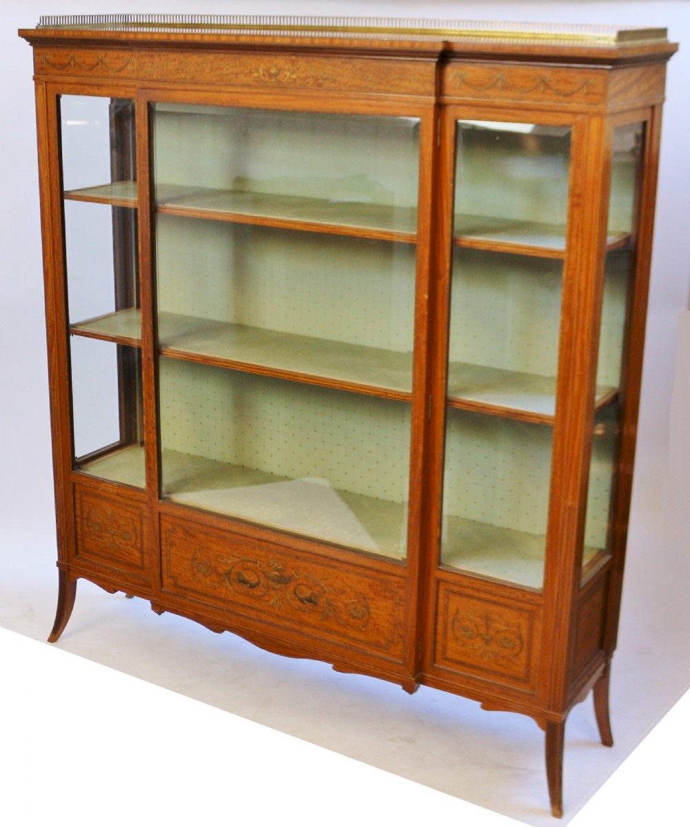 An Edwardian satinwood display cabinet featuring a breakfront design with glazed panels, floral marquetry inlay, and a brass top gallery. View IV