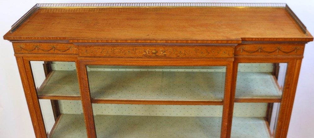 An Edwardian satinwood display cabinet featuring a breakfront design with glazed panels, floral marquetry inlay, and a brass top gallery. View VI