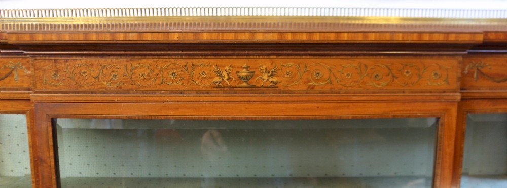 An Edwardian satinwood display cabinet featuring a breakfront design with glazed panels, floral marquetry inlay, and a brass top gallery. View X