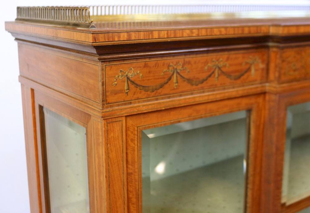 An Edwardian satinwood display cabinet featuring a breakfront design with glazed panels, floral marquetry inlay, and a brass top gallery. View XI