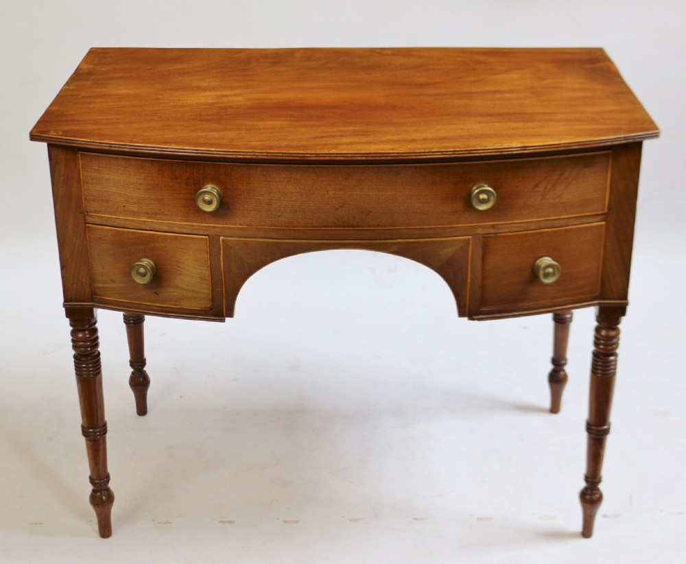 A Regency mahogany bowfront side table featuring three drawers with brass knob handles, an arched kneehole centre, and four turned legs.