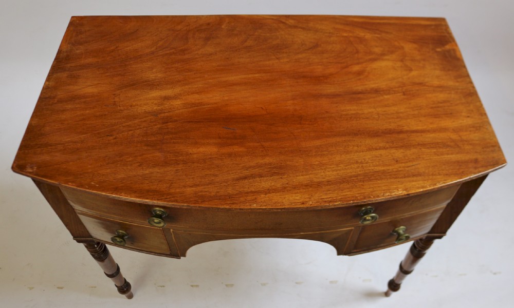 A Regency mahogany bowfront side table featuring three drawers with brass knob handles, an arched kneehole centre, and four turned legs. View VI