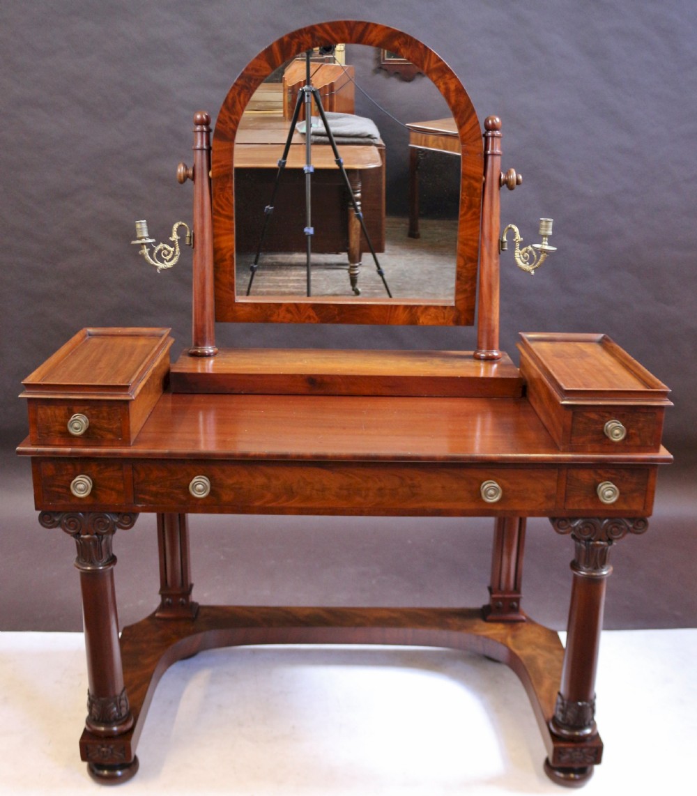 A William IV mahogany dressing table featuring an arched swivel mirror, brass sconces, upper drawers, and carved front columns on a plinth base.
