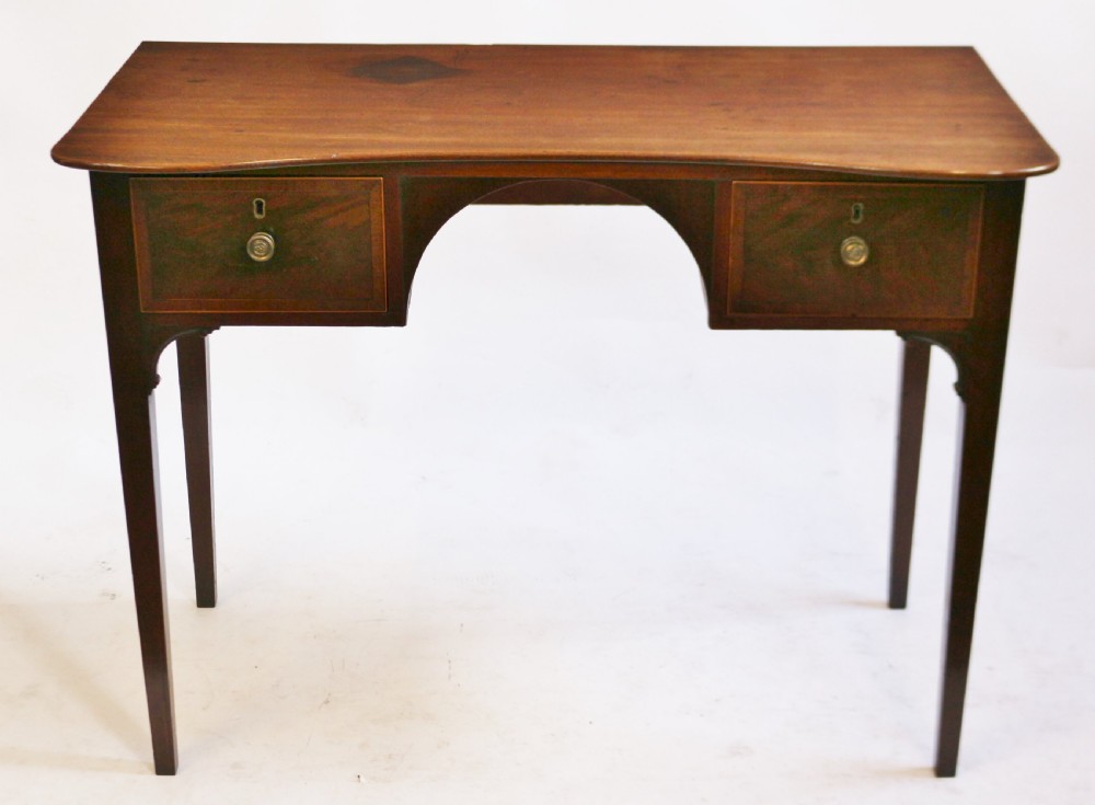 A George III mahogany kneehole table featuring a gently curved front, two string-inlaid drawers with brass handles, an arched central apron, and four square tapered legs.