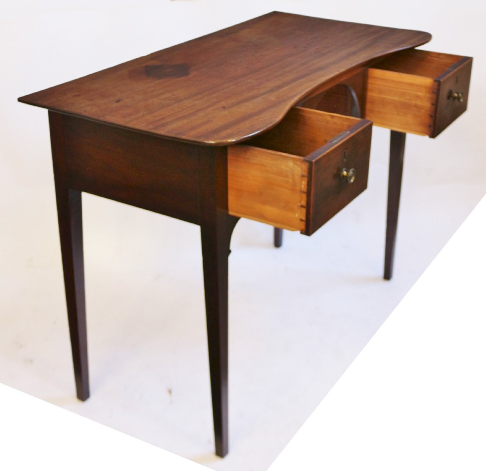 A George III mahogany kneehole table featuring a gently curved front, two string-inlaid drawers with brass handles, an arched central apron, and four square tapered legs. View IV