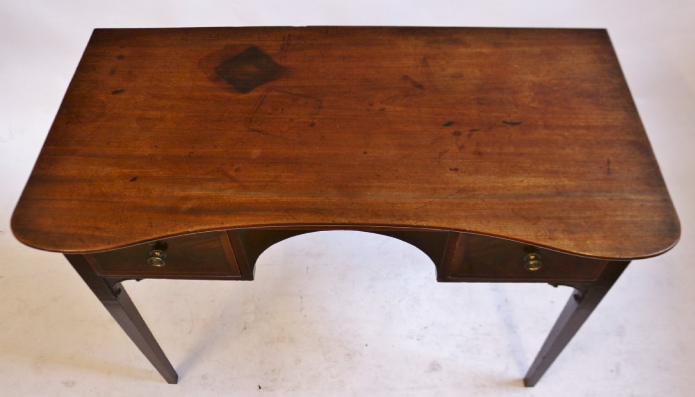 A George III mahogany kneehole table featuring a gently curved front, two string-inlaid drawers with brass handles, an arched central apron, and four square tapered legs. View V