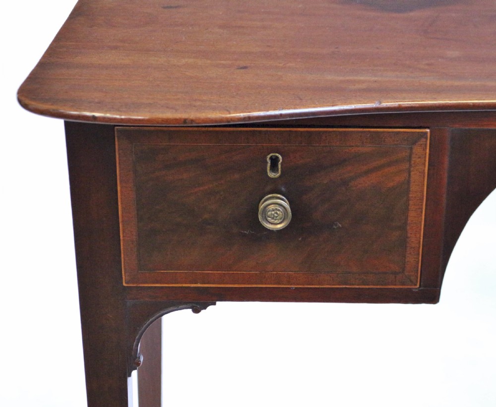 A George III mahogany kneehole table featuring a gently curved front, two string-inlaid drawers with brass handles, an arched central apron, and four square tapered legs. View VI