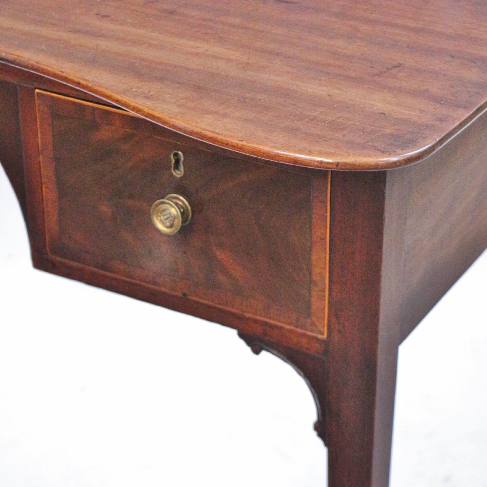A George III mahogany kneehole table featuring a gently curved front, two string-inlaid drawers with brass handles, an arched central apron, and four square tapered legs. View VII