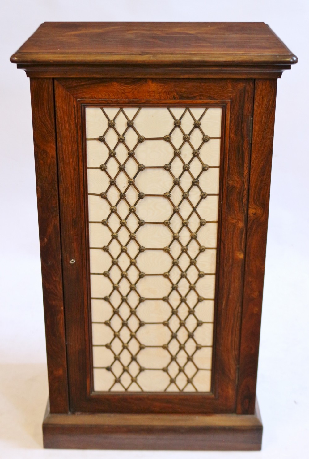 An antique William IV rosewood cupboard featuring a single door inset with a decorative brass lattice grille over a cream fabric backing.