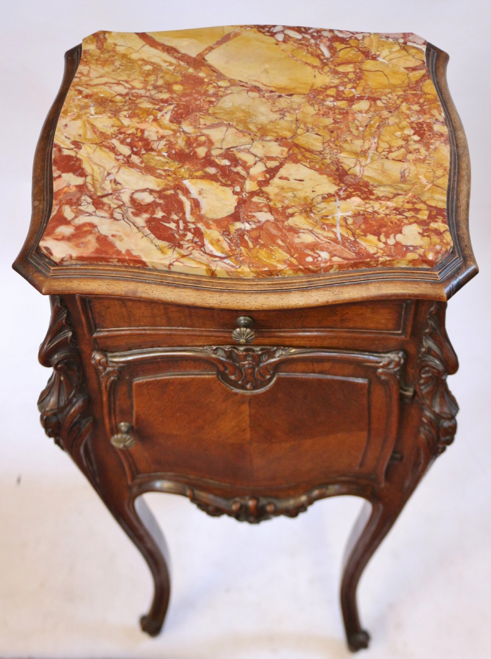 A 19th-century French walnut bedside cupboard featuring an inset variegated marble top, a top drawer, a quarter-veneered cupboard door, ornate rocaille carvings, and slender cabriole legs. View VI