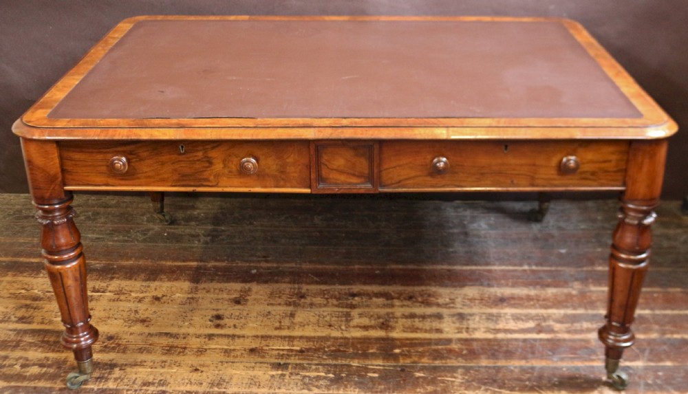 An early Victorian writing table in figured walnut featuring an inset brown leather top, four drawers with turned wooden knobs, and turned legs fitted with brass castors.