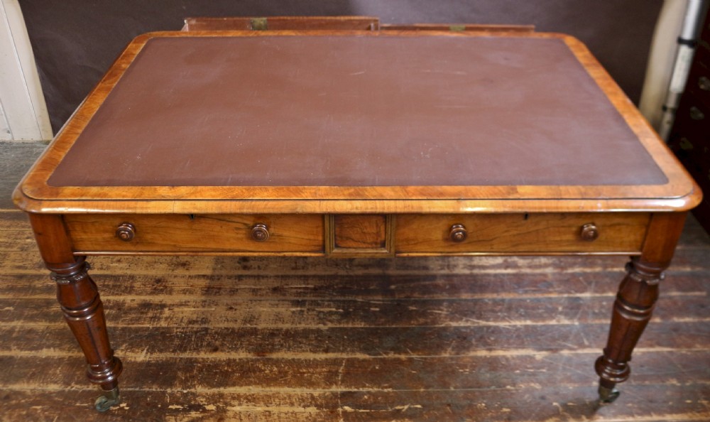 An early Victorian writing table in figured walnut featuring an inset brown leather top, four drawers with turned wooden knobs, and turned legs fitted with brass castors. View II