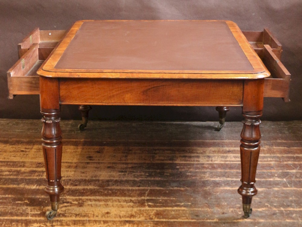 An early Victorian writing table in figured walnut featuring an inset brown leather top, four drawers with turned wooden knobs, and turned legs fitted with brass castors. View V