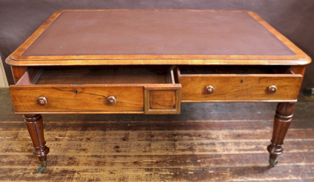 An early Victorian writing table in figured walnut featuring an inset brown leather top, four drawers with turned wooden knobs, and turned legs fitted with brass castors. View VI