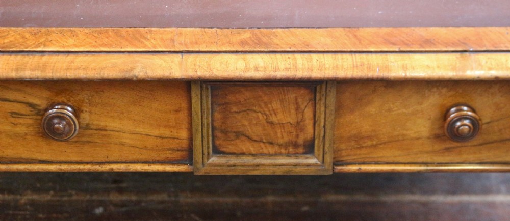 An early Victorian writing table in figured walnut featuring an inset brown leather top, four drawers with turned wooden knobs, and turned legs fitted with brass castors. View VIII