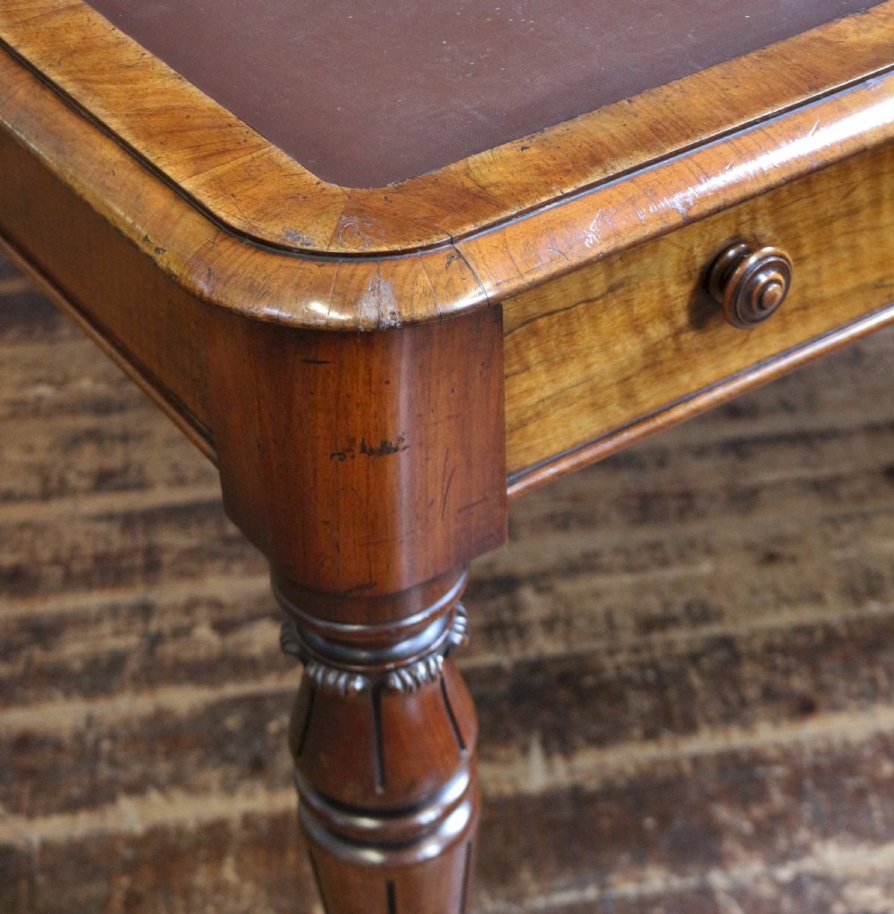 An early Victorian writing table in figured walnut featuring an inset brown leather top, four drawers with turned wooden knobs, and turned legs fitted with brass castors. View IX