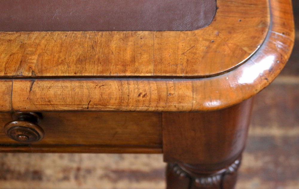 An early Victorian writing table in figured walnut featuring an inset brown leather top, four drawers with turned wooden knobs, and turned legs fitted with brass castors. View XI