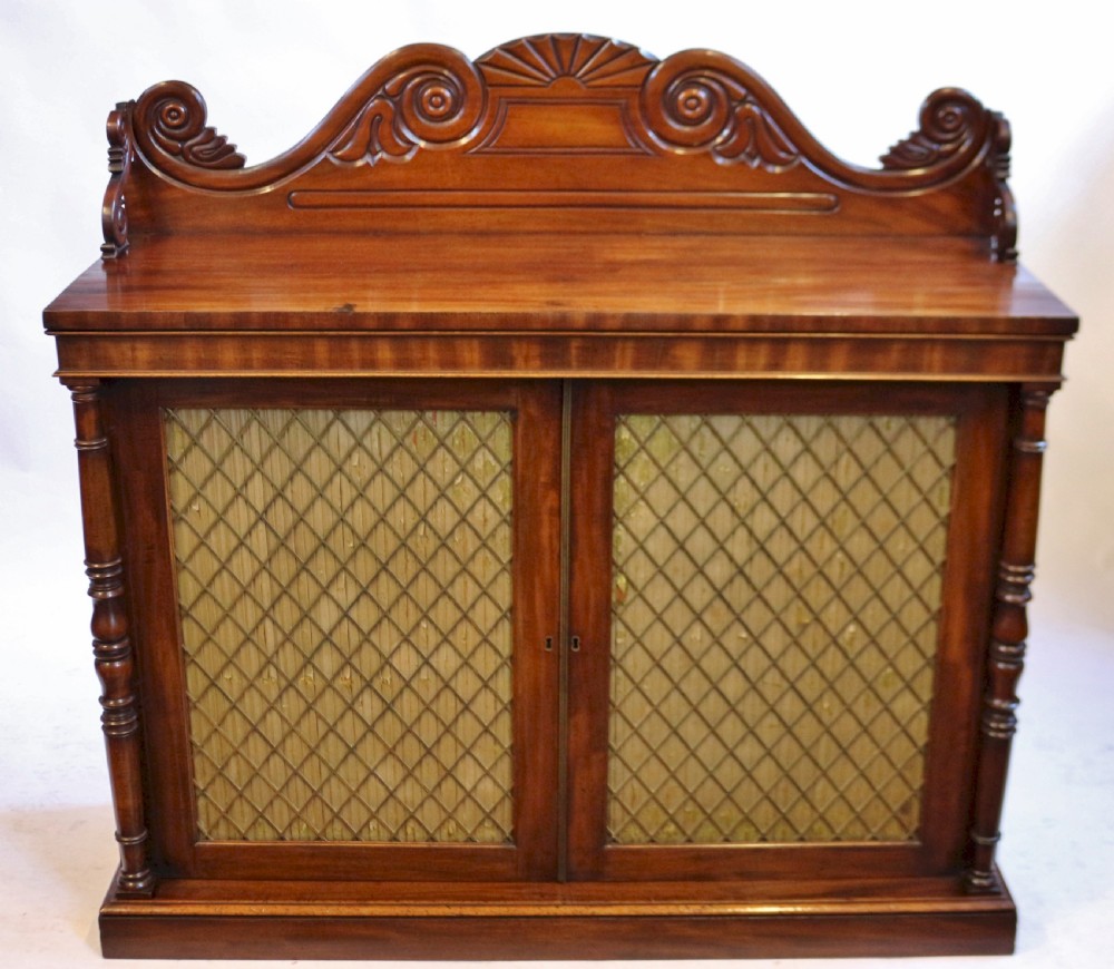 A William IV flame mahogany two-door side cabinet featuring a carved scroll upstand, turned columns, and brass wire mesh panels over pleated lining.