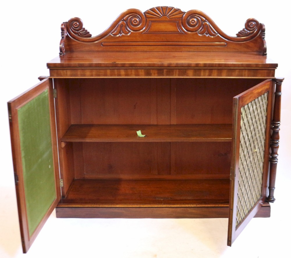 A William IV flame mahogany two-door side cabinet featuring a carved scroll upstand, turned columns, and brass wire mesh panels over pleated lining. View II