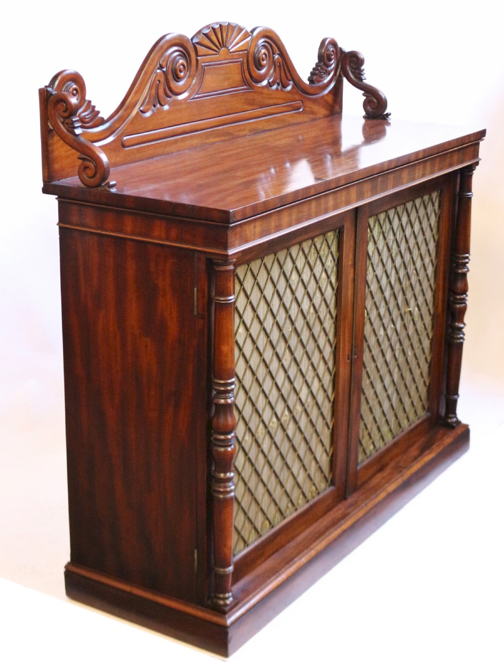 A William IV flame mahogany two-door side cabinet featuring a carved scroll upstand, turned columns, and brass wire mesh panels over pleated lining. View III