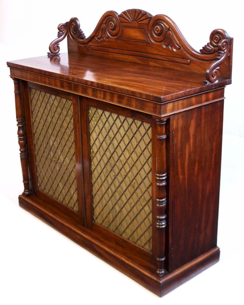 A William IV flame mahogany two-door side cabinet featuring a carved scroll upstand, turned columns, and brass wire mesh panels over pleated lining. View IV