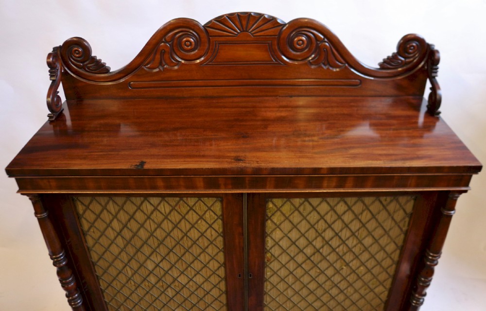 A William IV flame mahogany two-door side cabinet featuring a carved scroll upstand, turned columns, and brass wire mesh panels over pleated lining. View V