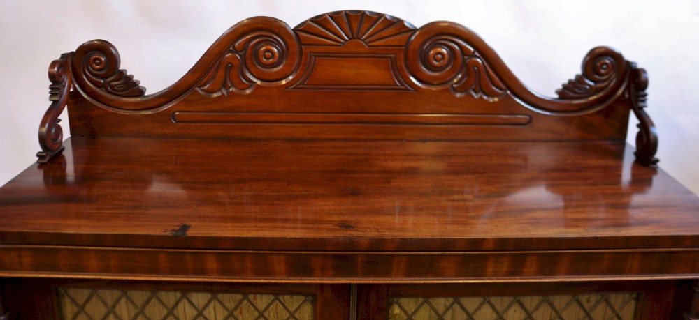 A William IV flame mahogany two-door side cabinet featuring a carved scroll upstand, turned columns, and brass wire mesh panels over pleated lining. View VI