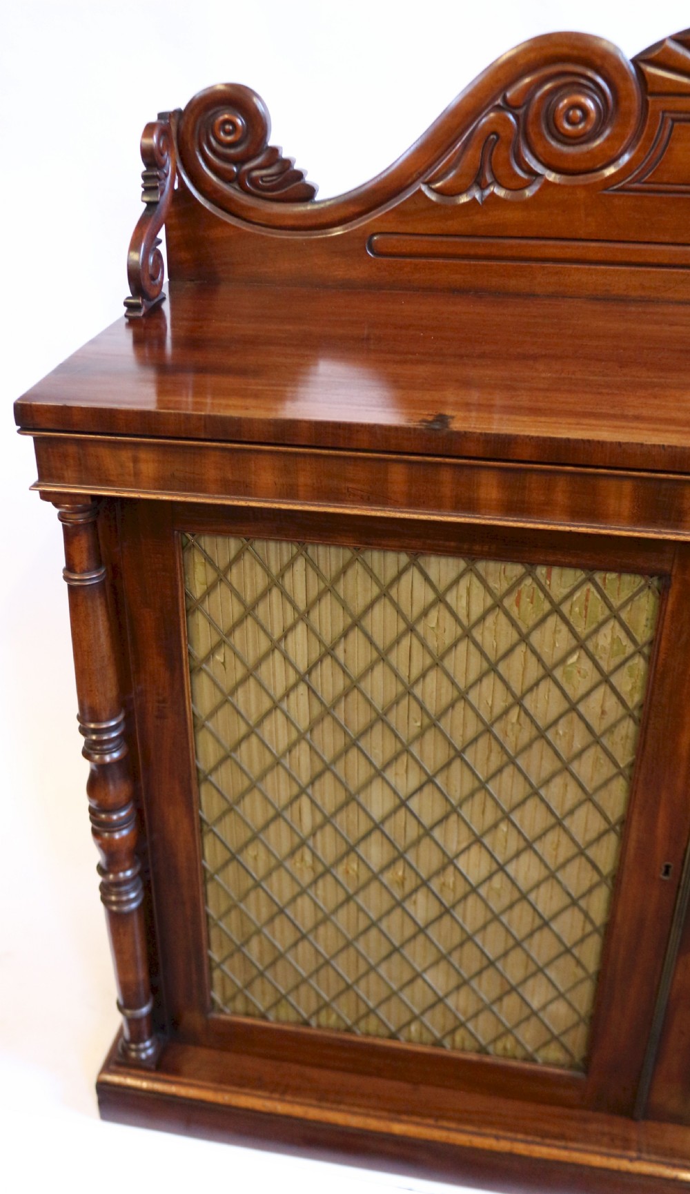 A William IV flame mahogany two-door side cabinet featuring a carved scroll upstand, turned columns, and brass wire mesh panels over pleated lining. View VII