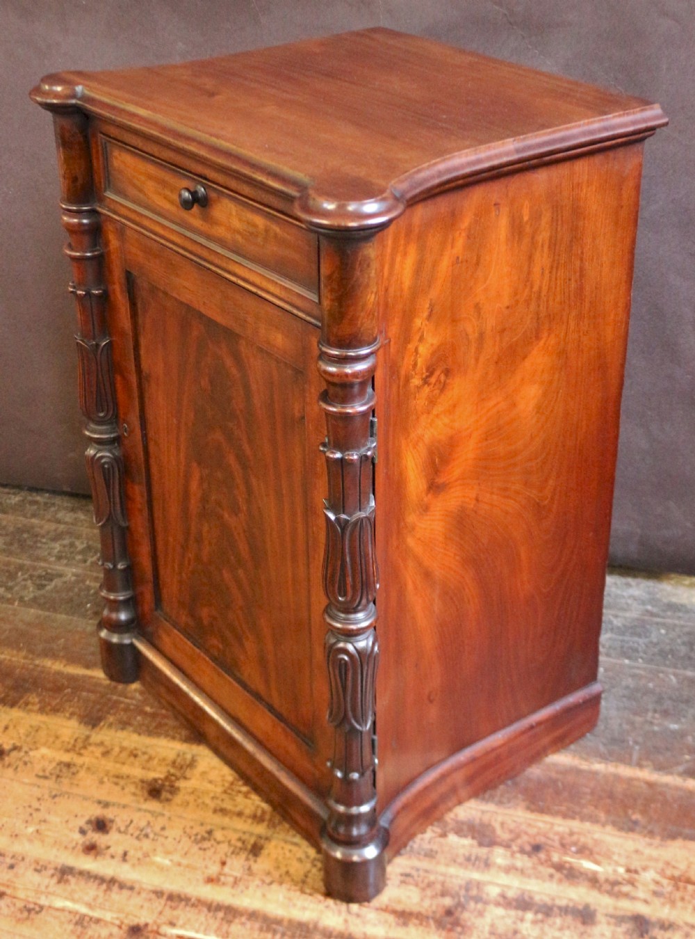 A William IV flame mahogany single-door bedside cupboard featuring a top drawer, carved acanthus leaf side columns, and a plinth base. View II