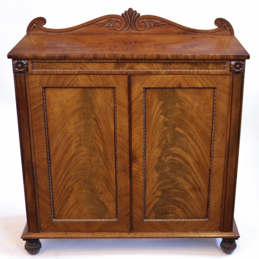 A William IV mahogany two-door side cupboard featuring flame mahogany door panels, a carved scroll backboard, and turned carved feet.