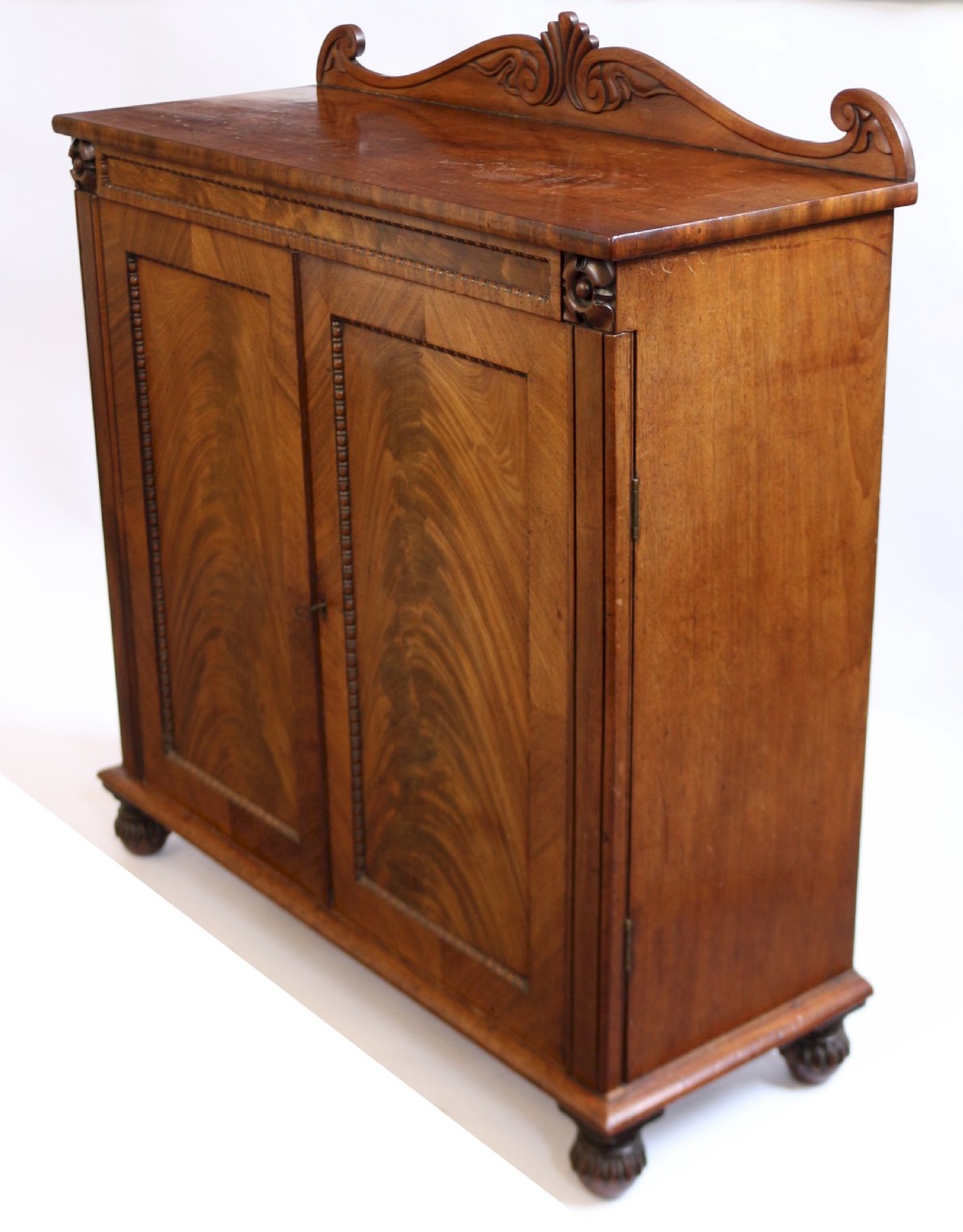 A William IV mahogany two-door side cupboard featuring flame mahogany door panels, a carved scroll backboard, and turned carved feet. View IV