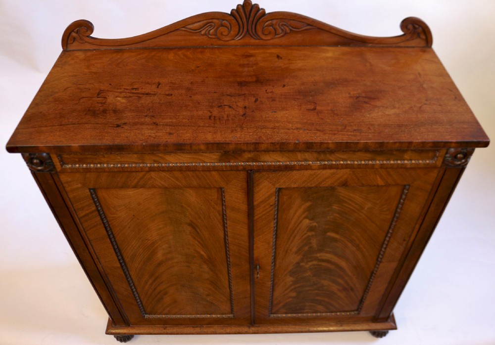 A William IV mahogany two-door side cupboard featuring flame mahogany door panels, a carved scroll backboard, and turned carved feet. View V