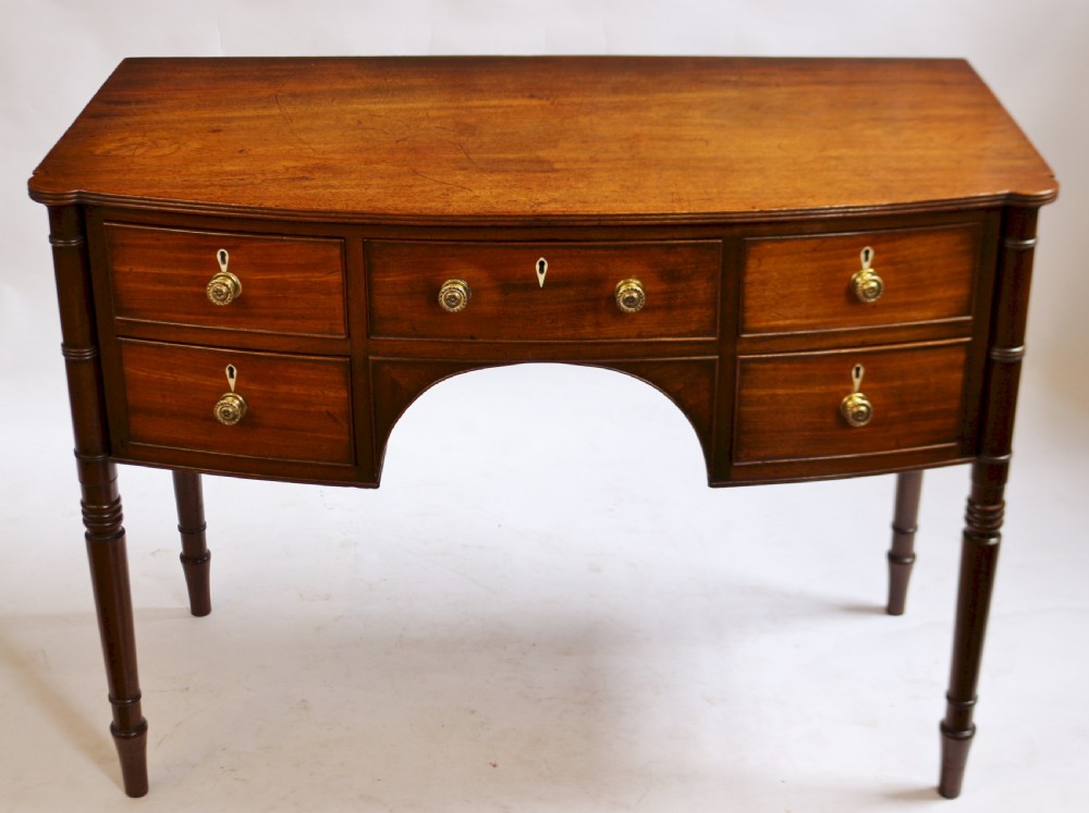A George IV mahogany bowfront kneehole table with five drawers, brass knobs, teardrop escutcheons, and turned legs.