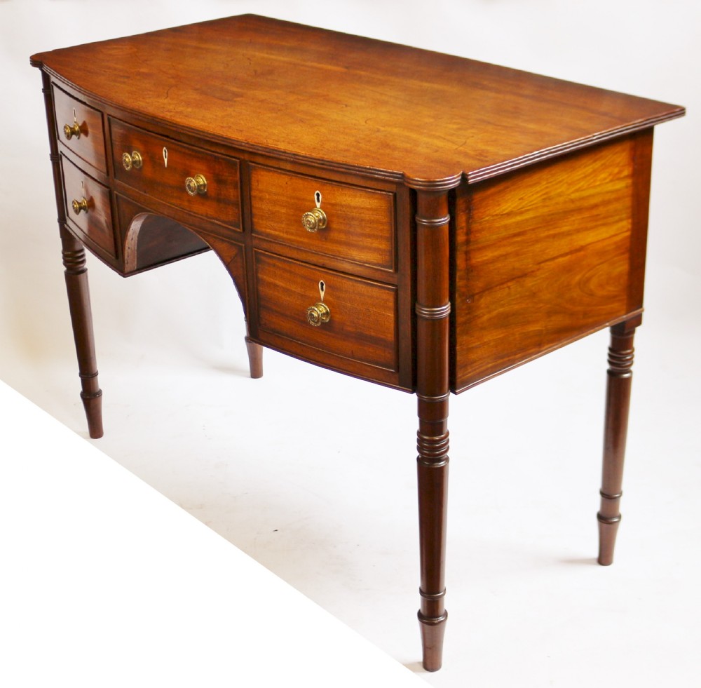 A George IV mahogany bowfront kneehole table with five drawers, brass knobs, teardrop escutcheons, and turned legs. View IV
