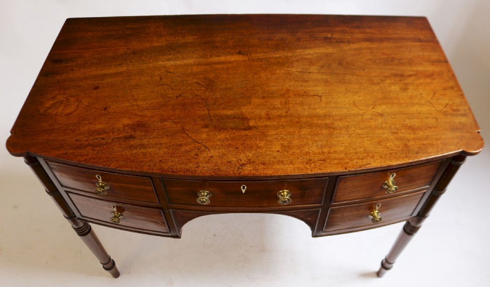 A George IV mahogany bowfront kneehole table with five drawers, brass knobs, teardrop escutcheons, and turned legs. View V