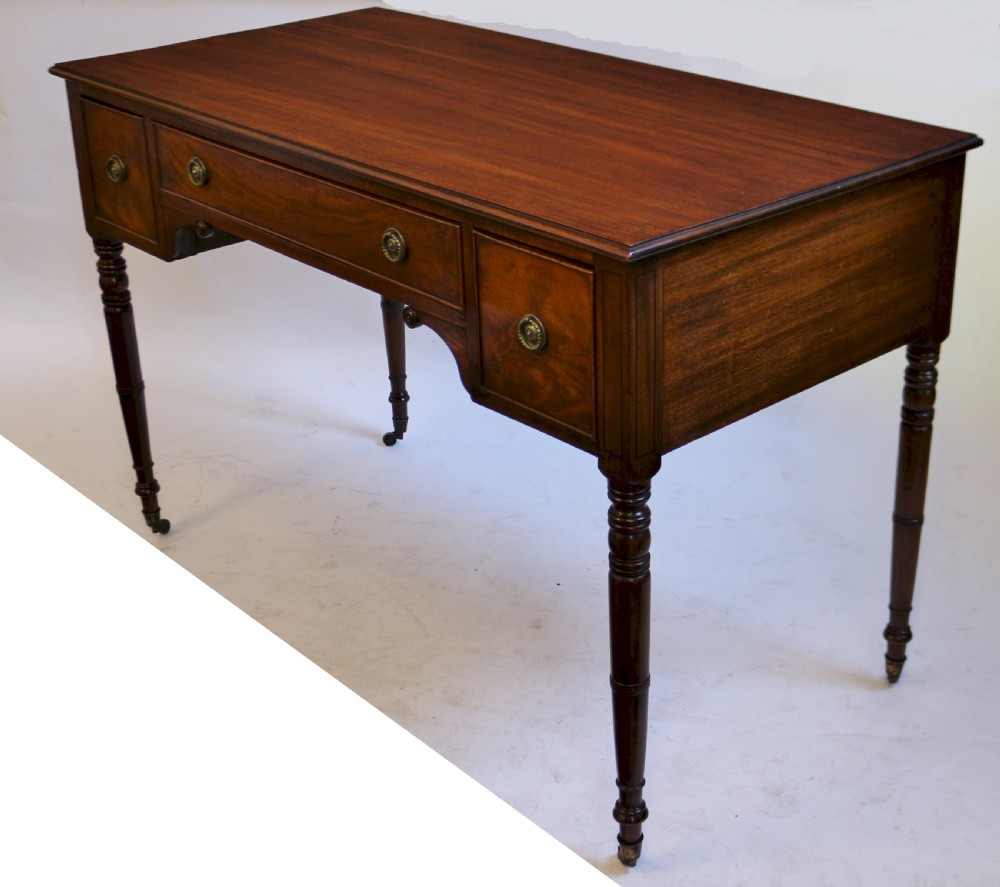 A George IV mahogany kneehole writing table with three drawers, turned legs, brass rosette handles, and brass castors. View III