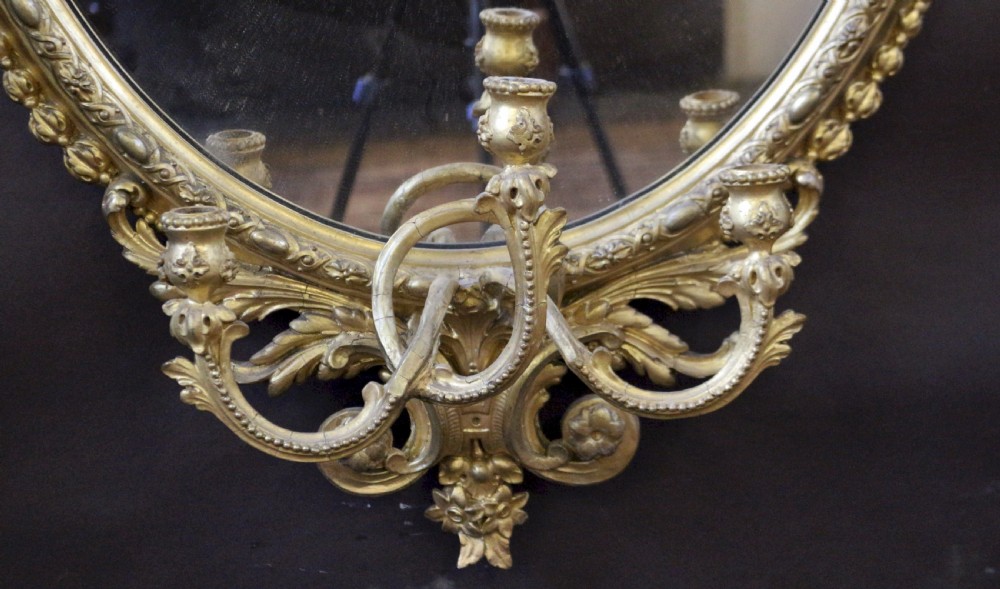 An oval giltwood wall mirror featuring a laurel leaf crest with a central urn motif and integrated candle sconces at the base. View II