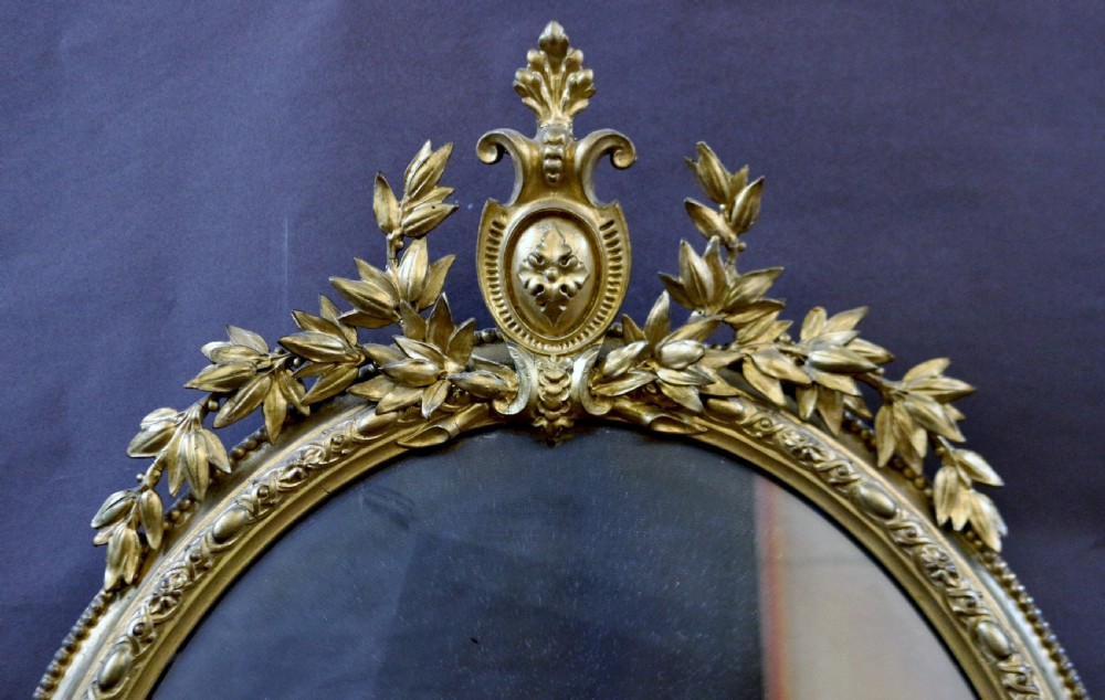 An oval giltwood wall mirror featuring a laurel leaf crest with a central urn motif and integrated candle sconces at the base. View III