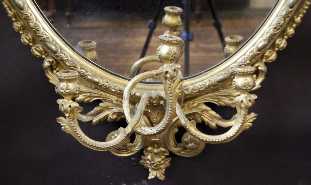 An oval giltwood wall mirror featuring a laurel leaf crest with a central urn motif and integrated candle sconces at the base. View VII