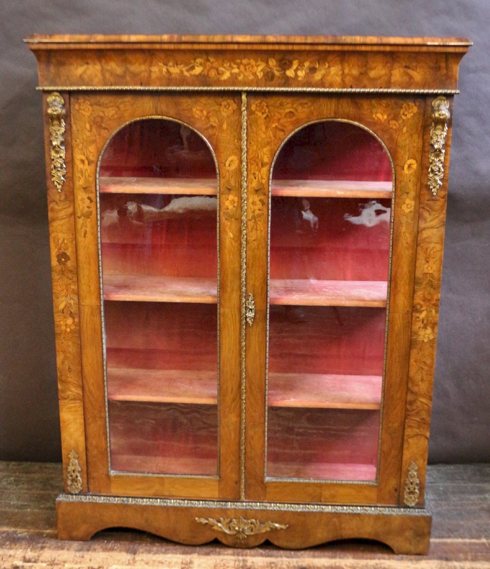 A Victorian burr walnut pier cabinet featuring floral marquetry inlays, gilt ormolu mounts, and double arched glass doors revealing shelves lined in red fabric.
